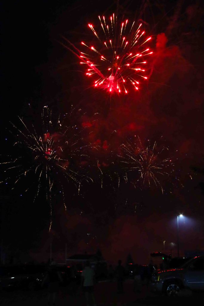 A display of red fireworks lighting up the night sky, with streaks of light and smoke in the background. Silhouetted figures are seen below, possibly watching the display.