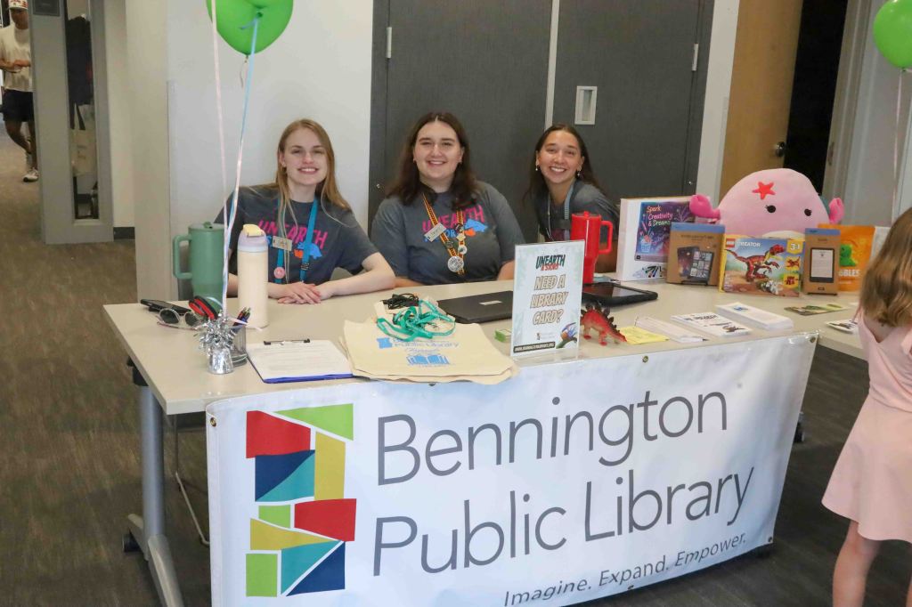 Three young women smiling behind a table with a Bennington Public Library banner, showcasing library materials and registration information.