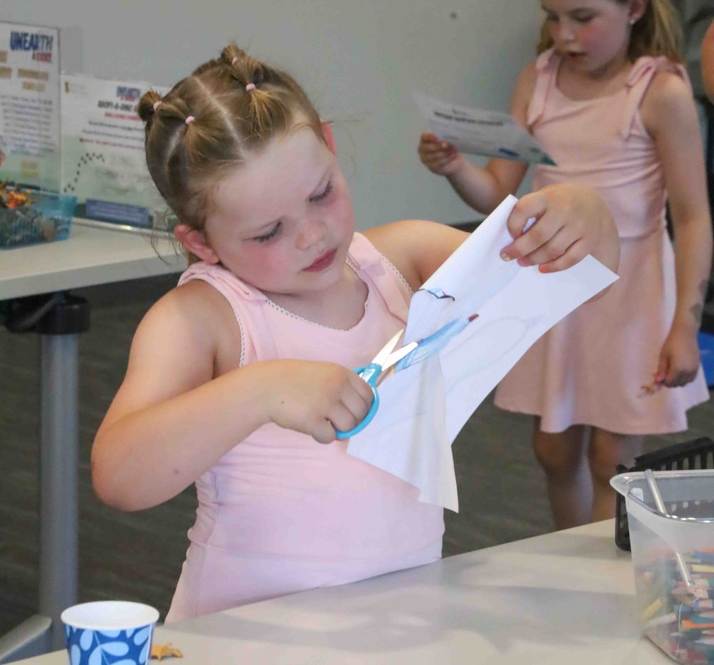 A young girl in a pink dress is cutting paper with blue scissors, focusing intently on her task. Another girl in the background is holding a piece of paper.
