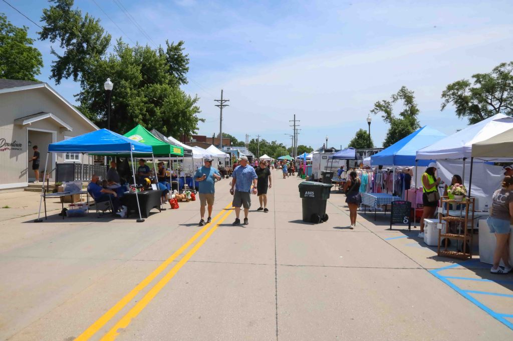 A busy outdoor market street with colorful tents, vendors selling various items, and people walking between them on a sunny day.