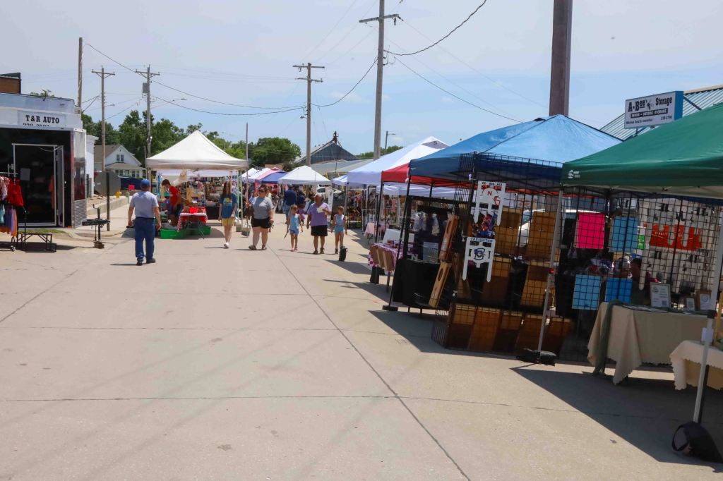 A bustling street market scene with various vendor tents in different colors, people shopping and browsing, and utility poles in the background.