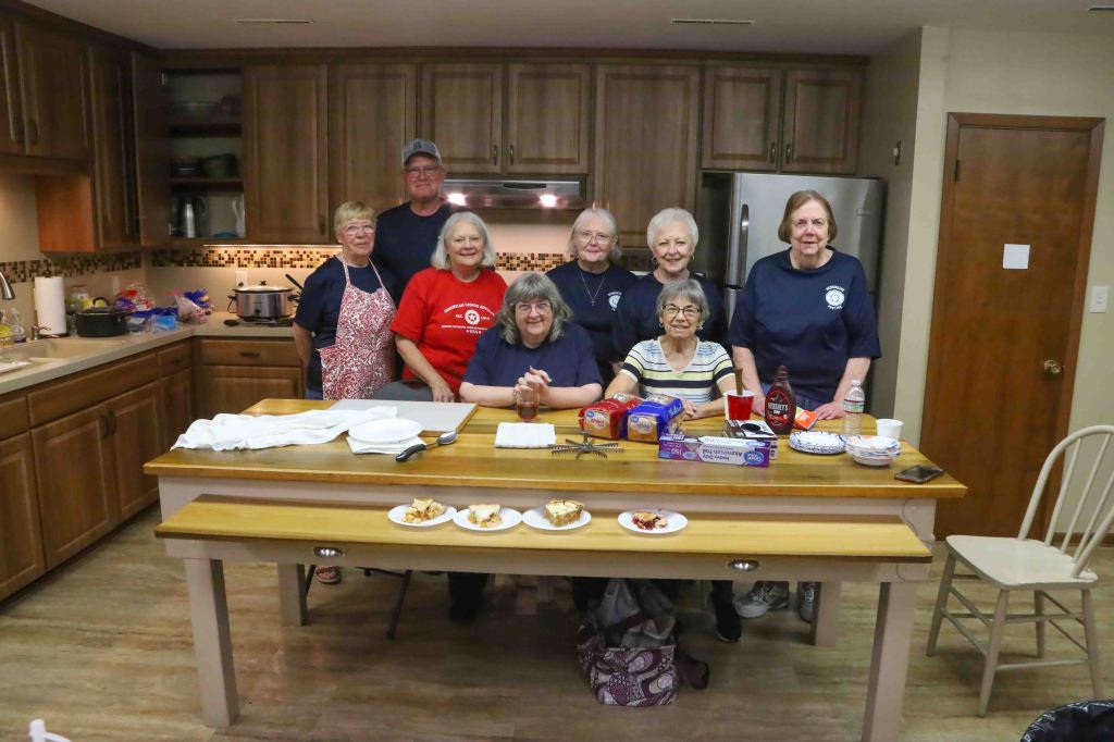 Group of people gathered in a kitchen, smiling and posing for a photo. There are various food items on the table, including desserts and snacks.
