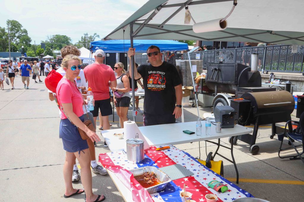 A food vendor at a festival with a barbecue setup and a man wearing a black t-shirt standing behind a table. A woman in a pink shirt is in the foreground, while people browse around in the background. The table is decorated with a star-patterned tablecloth.