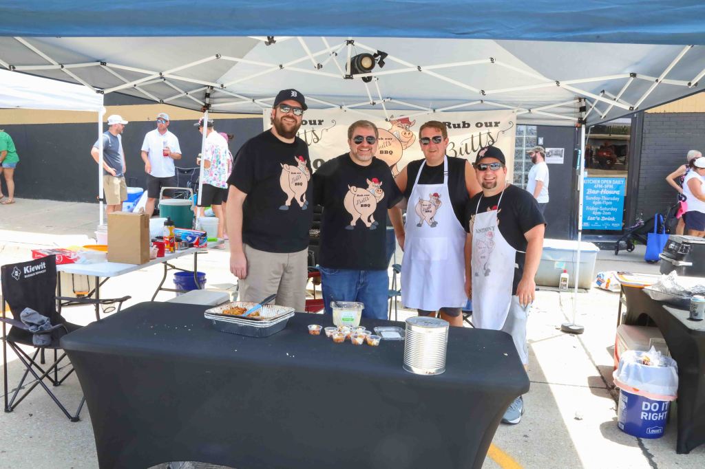 Four men standing behind a table at a barbecue event, wearing black shirts with a humorous pig design and aprons. They are smiling and posing for the camera, with various food items on the table and a crowd in the background.