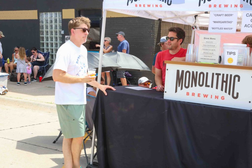 A man in a white t-shirt holding a drink at a brewing booth labeled 'Monolithic Brewing'. Another man in a red shirt is behind the counter, attending to customers. People are seated in the background, enjoying the event outdoors.