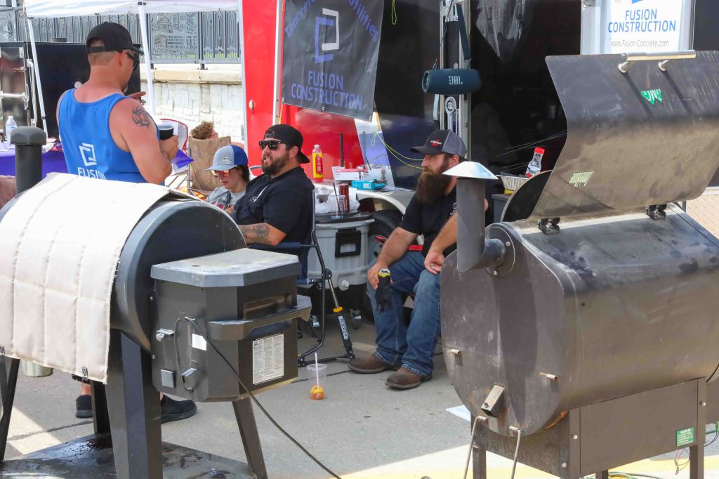 A group of men relaxing at a construction event, with one man holding a drink and two others seated nearby in front of a food trailer. Smokers are visible in the foreground.