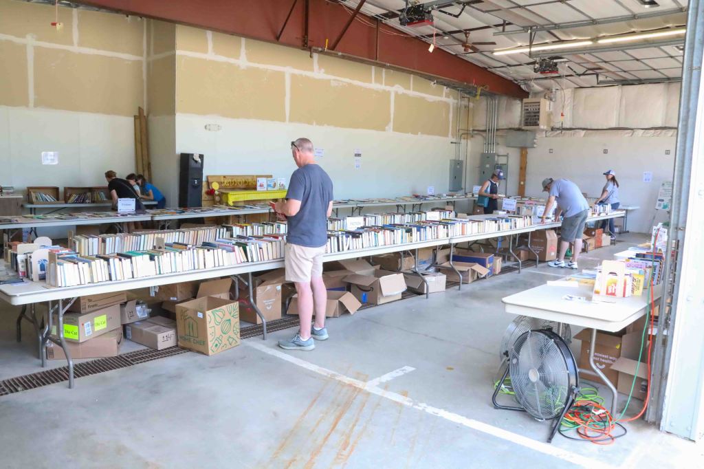A large indoor space with multiple tables arranged in rows, displaying numerous books and boxes. Several people are browsing through the books, while one individual is checking their phone. The ceiling has exposed beams, and there are various electrical installations visible on the walls.