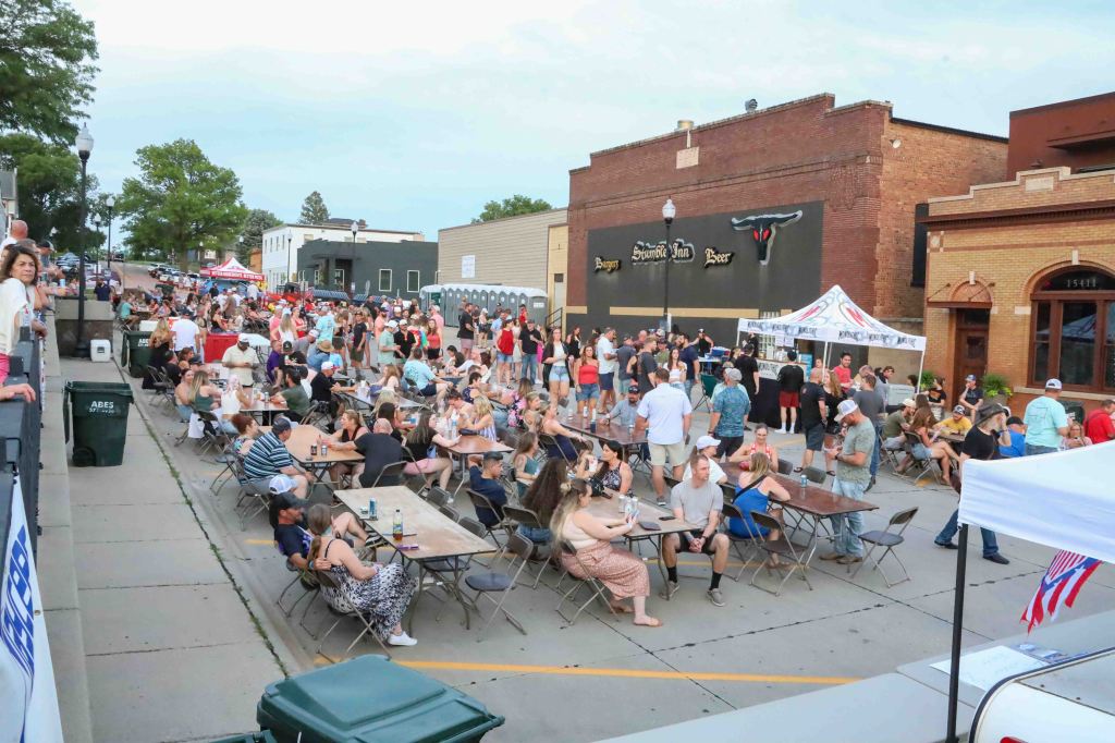 A lively outdoor gathering with a large crowd sitting at tables, enjoying food and drinks on a street lined with shops and trees. The scene captures people socializing and participating in a fun atmosphere during an event.