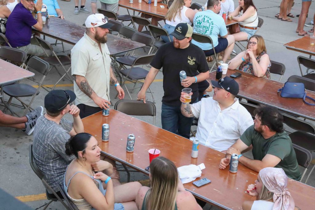 A lively outdoor gathering with people sitting at tables, some interacting and enjoying drinks. A few individuals are engaged in conversation, with beer cans and cups on the table.
