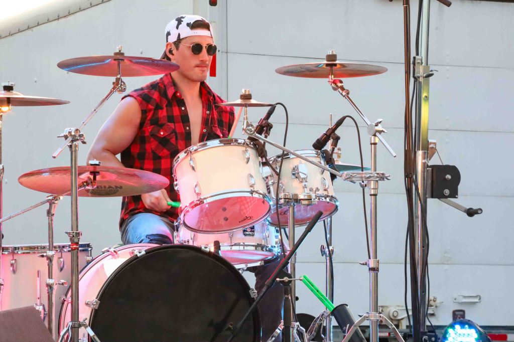 A drummer playing on stage, wearing sunglasses and a red plaid shirt, with a white drum set and cymbals in the background.