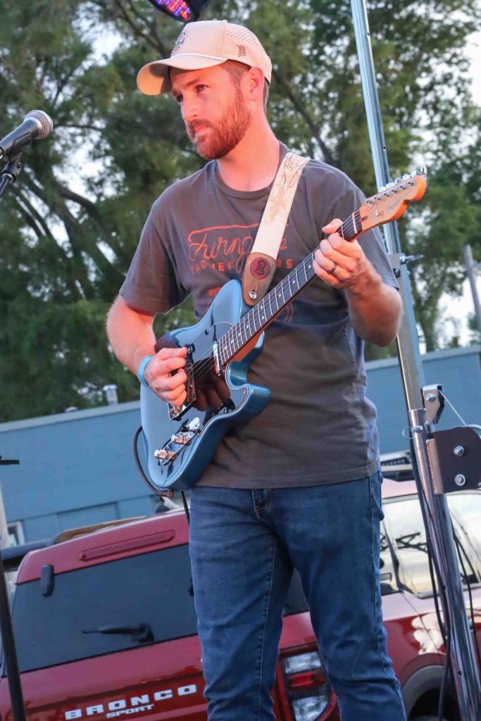 A musician playing a blue electric guitar on stage, wearing a cap and a t-shirt. The background shows a red vehicle parked nearby.