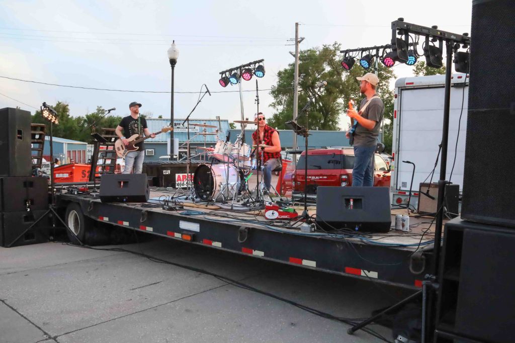A live band performing on a stage set on a trailer, with three musicians playing guitar and drums, under colorful stage lights. In the background, vehicles and buildings are visible.