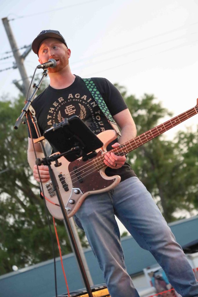 A musician performing on stage with a bass guitar, wearing a black cap and a t-shirt. The background features trees and power lines, with a setting sun.