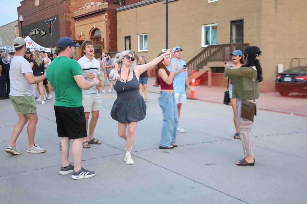 A group of people enjoying a lively outdoor event, dancing and interacting on a street, with buildings and a crowd in the background.