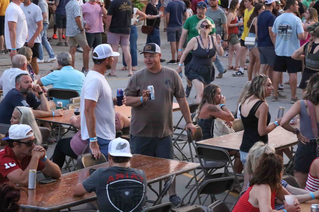 A lively outdoor event scene with groups of people socializing, some sitting at tables, while others are walking around. Attendees hold drinks and wear casual summer attire, showcasing a festive atmosphere.