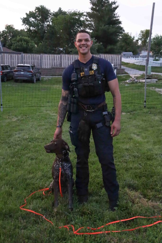 A smiling police officer in tactical gear stands on grass with a dog beside him, both appearing relaxed in an outdoor setting.