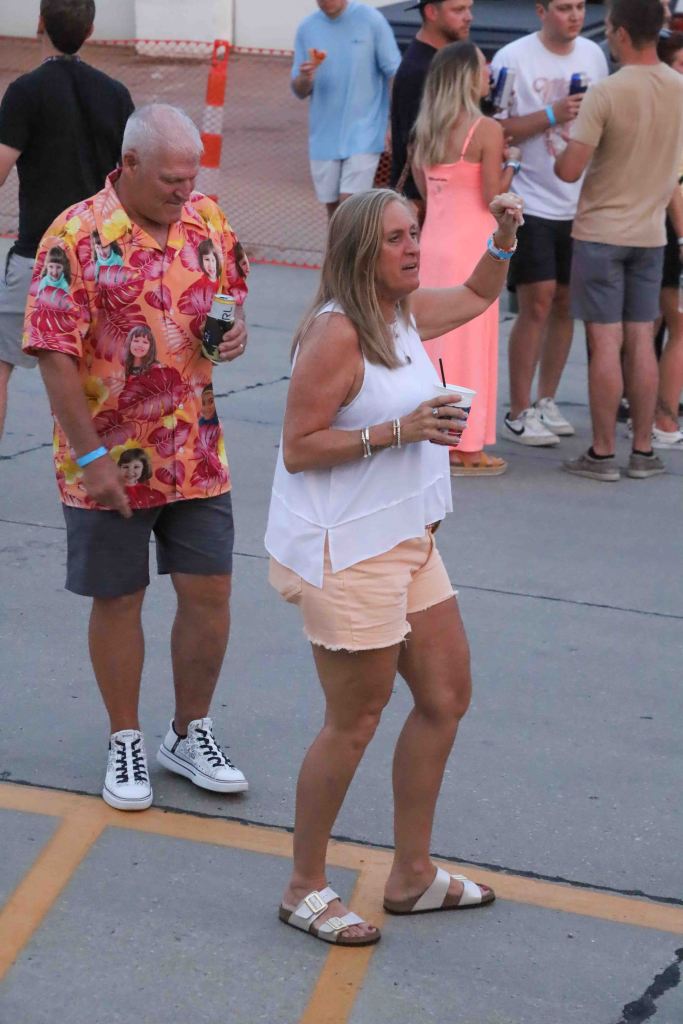 A woman in a white top and shorts drinks from a cup while dancing, alongside a man in a colorful patterned shirt and shorts, in a lively outdoor event.
