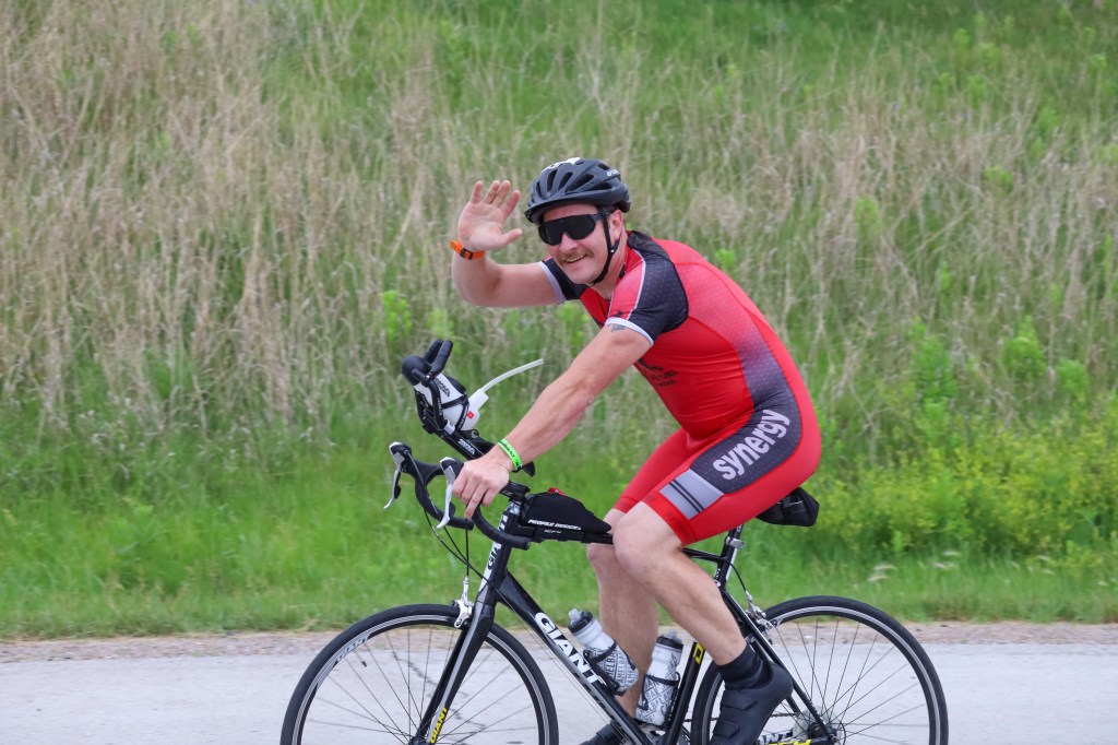 A cyclist wearing a red racing suit waves while riding a bike along a grassy path.