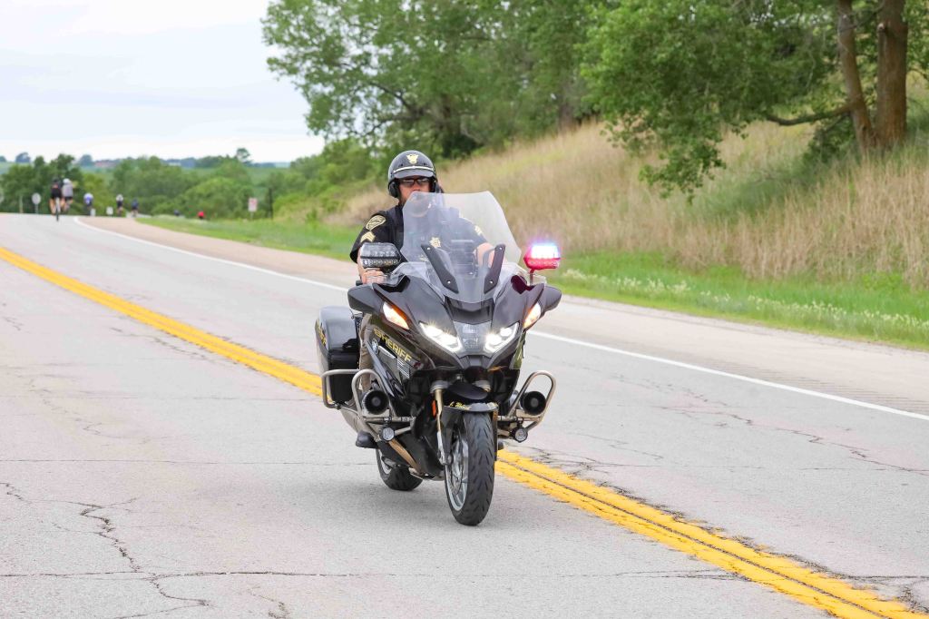 A police officer riding a black motorcycle on a rural road, with lush greenery in the background.