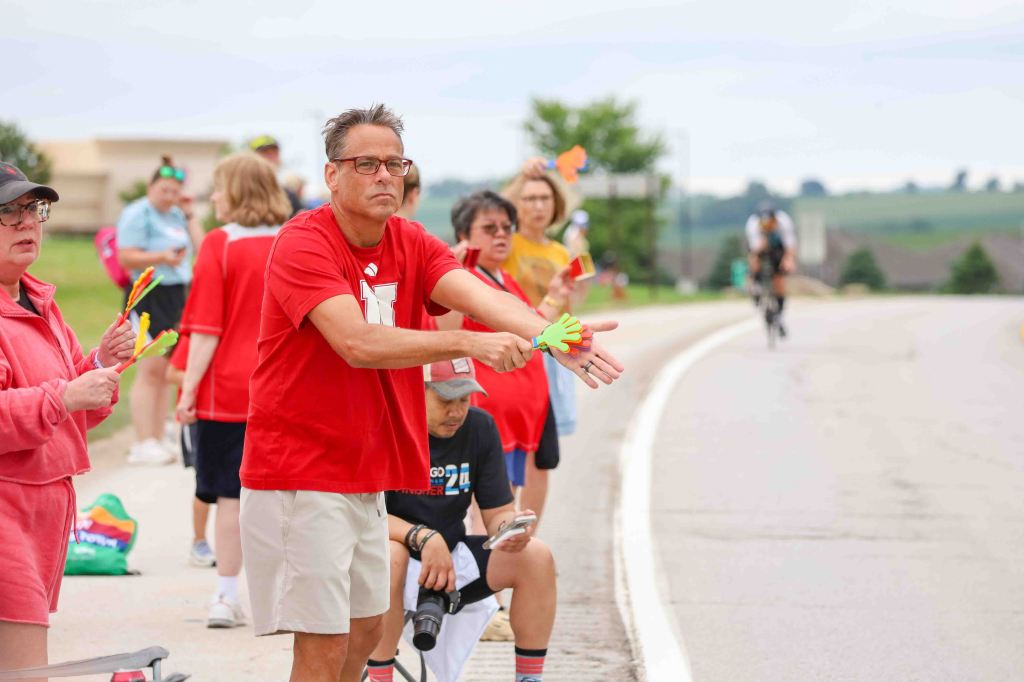 A group of supporters cheer on cyclists from the roadside, wearing colorful clothing and holding accessories like hand-shaped clappers. One man in a red shirt and glasses is prominently waving his hands in encouragement.
