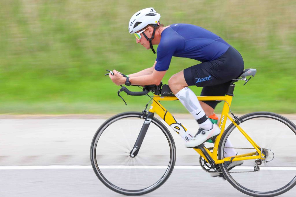 A cyclist in a blue shirt and black shorts riding a yellow road bike on a paved road, captured in motion with a blurred background.