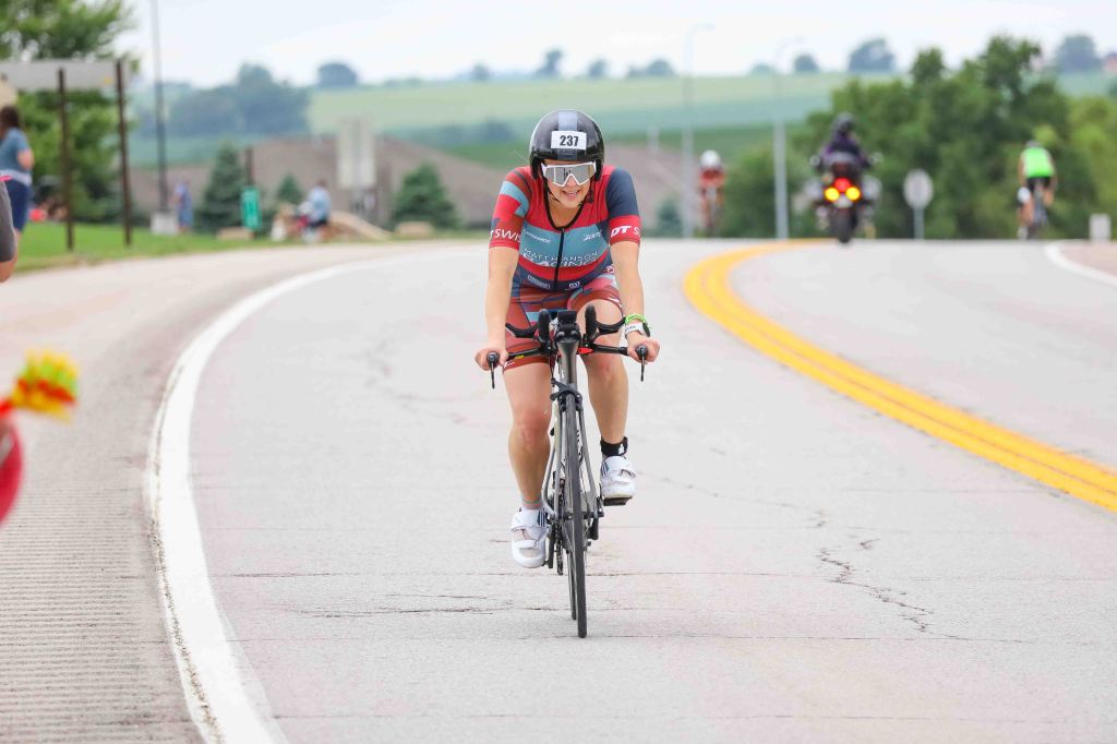 A cyclist wearing a helmet and a blue and red cycling outfit rides on a winding road, surrounded by greenery and other cyclists in the background.
