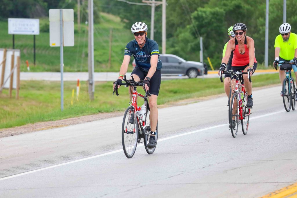 A group of cyclists riding on a road, with two people in the foreground smiling and pedaling, wearing helmets and cycling attire, surrounded by greenery and road signs in the background.