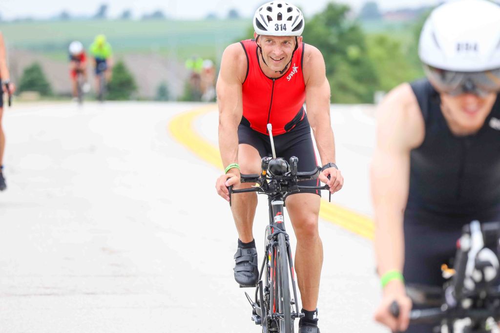 A male triathlete, wearing a red sleeveless suit and a helmet, smiles as he rides a bicycle on a road during a competition. Other cyclists are visible in the background.