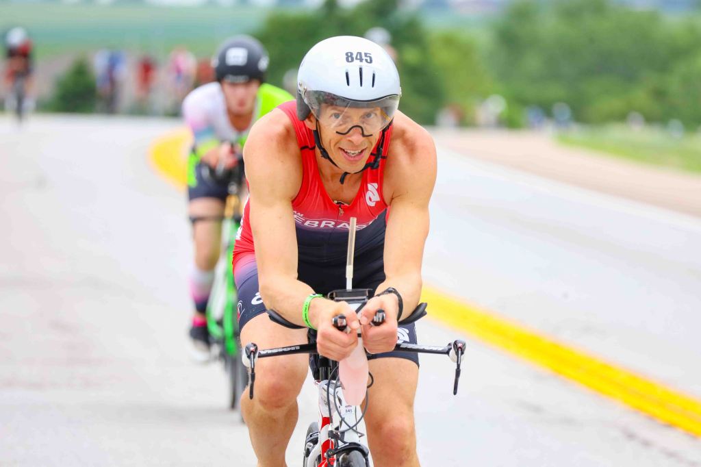 A competitive cyclist in a red triathlon suit races on a road, with a focused expression. Other cyclists are visible in the background, suggesting a triathlon event.