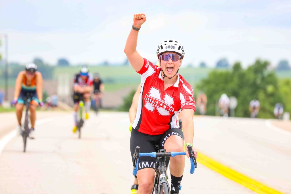 A cyclist in a red jersey with 'HUSKERS' celebrates with a raised fist while riding on a rural road during a race, with other cyclists visible in the background.
