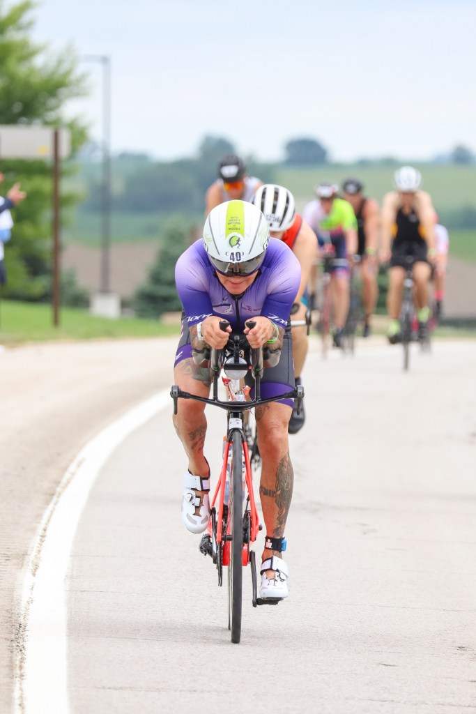 A group of cyclists participating in a triathlon on a road, with one athlete in the foreground riding a bright orange bike and wearing a purple cycling suit.