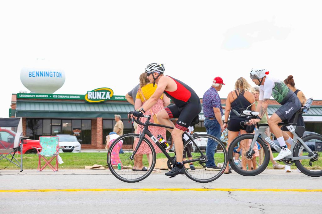 Two cyclists participating in a race, riding past a restaurant and water tower labeled 'Bennington'. Spectators are gathered along the side of the road.