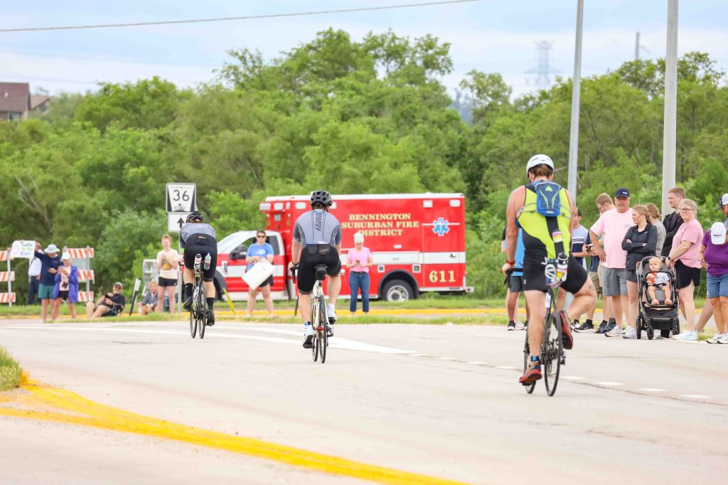 Two cyclists riding along a road with spectators watching nearby, including a family with a stroller, and an emergency vehicle in the background.