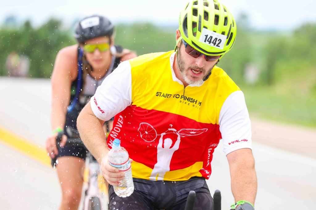 A male cyclist in a colorful jersey rides along a wet road, holding a water bottle. Another cyclist is visible in the background.