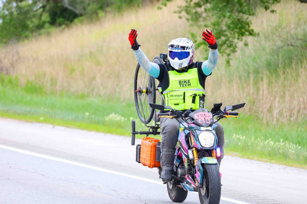 A motorcycle rider wearing a helmet and safety vest stands on their bike with arms raised. They are holding a bicycle wheel on their back and have a tool box attached to the bike, set against a grassy roadside background.