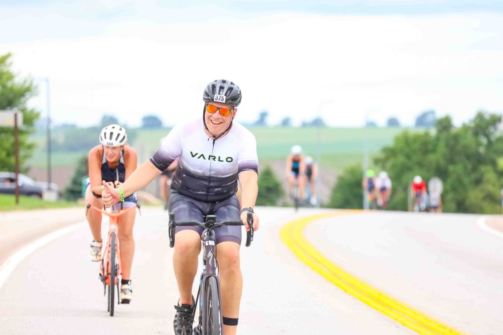 A group of cyclists riding on a winding road, with a smiling male cyclist in the foreground wearing a VARLO jersey and sunglasses, and a female cyclist in the background.