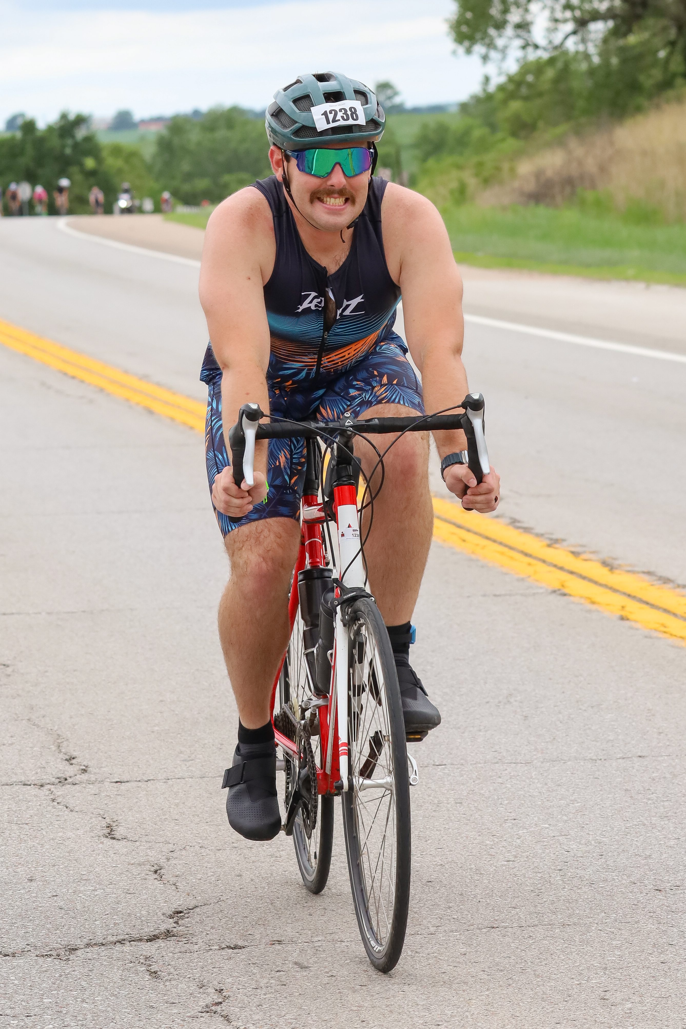 A male cyclist with a mustache rides a red bicycle on a country road, wearing a helmet and sunglasses, and sporting a colorful racing outfit.