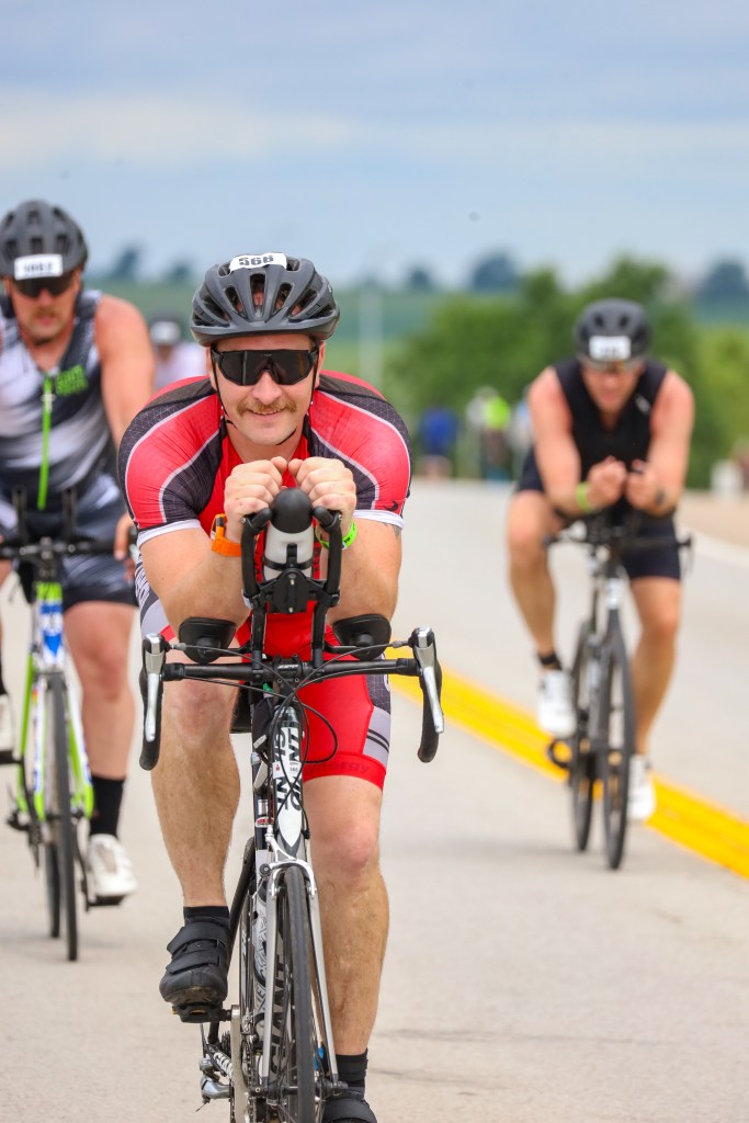 A group of triathletes cycling on a rural road. The focus is on a male cyclist wearing a red and black racing outfit, sunglasses, and a helmet, as he leans forward on his bike. Two other cyclists are visible in the background.
