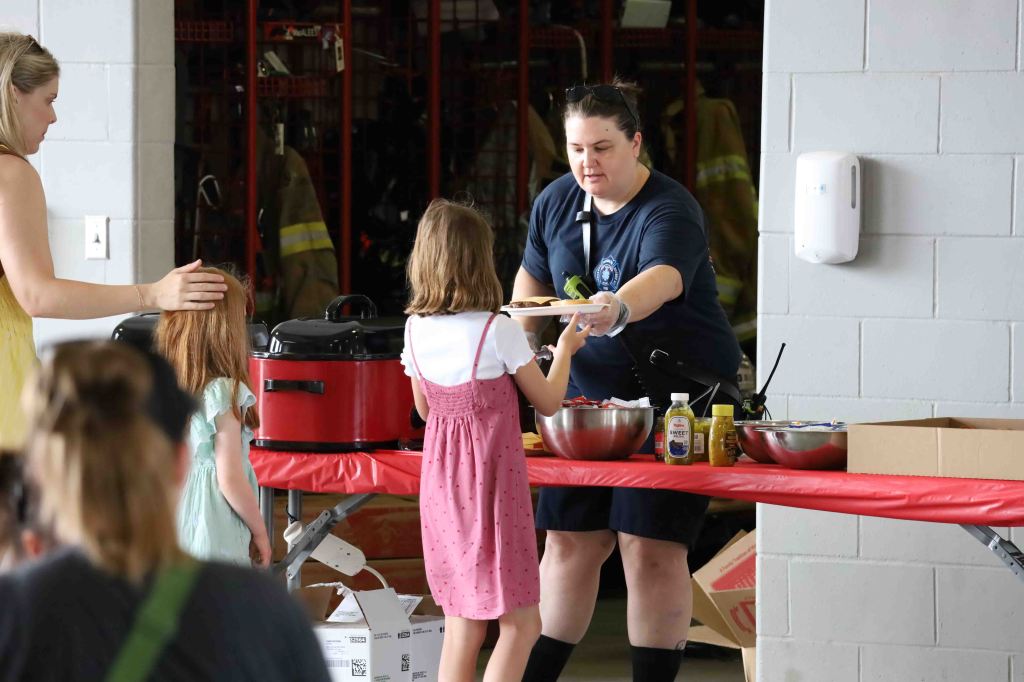 A woman serving food to a young girl at a table in a community hall, with other children waiting in line.