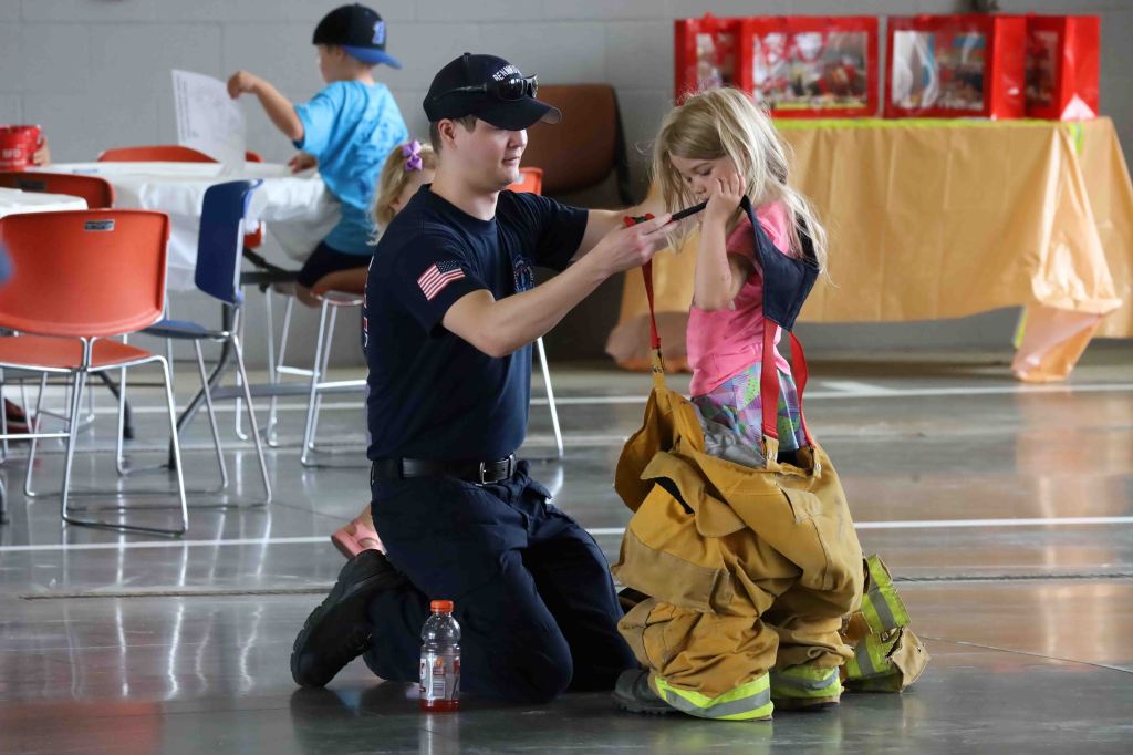 A firefighter helps a young child try on oversized firefighting gear in a community setting.