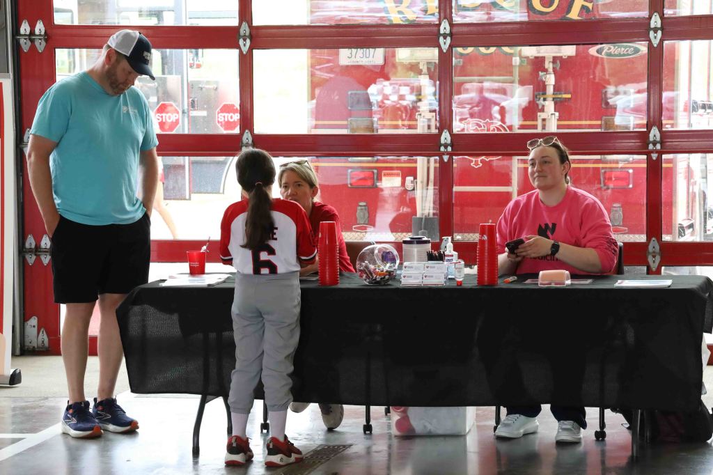 A girl in a baseball uniform stands at a table with two adults, one in a pink shirt and the other in a blue shirt, inside a fire station with a fire truck visible in the background.