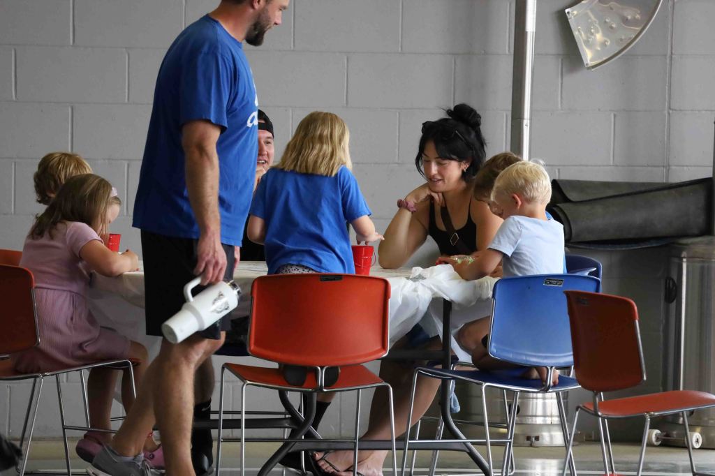 A group of children and adults sitting around a table, enjoying a meal together in a casual setting. Two adults are interacting with the kids, who are engaged with their food.