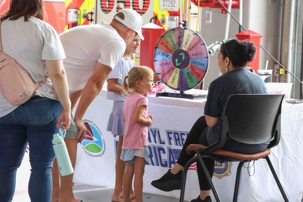 A family interacts at a community event, with a little girl excitedly approaching a wheel of prizes while an adult sits behind a table.