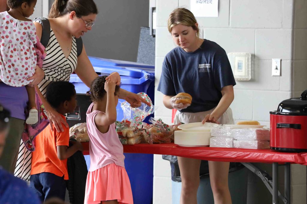 A woman serving food at a table with children nearby, one holding her head while another reaches out for a snack.