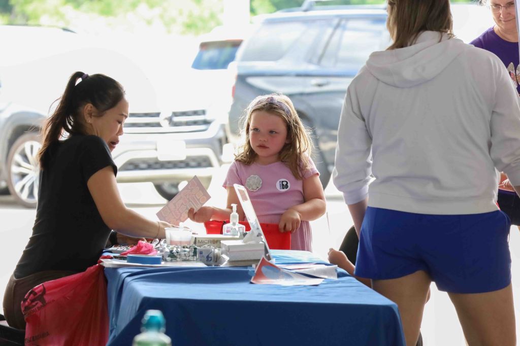 A young girl in a pink shirt interacts with an adult at a table, which is covered in supplies. The adult is handing over a paper while another person stands nearby, and cars are visible in the background.