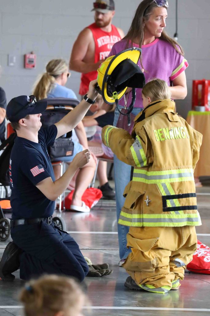 A firefighter kneels to assist a child wearing oversized firefighter gear, including a jacket and helmet, in a community event setting.