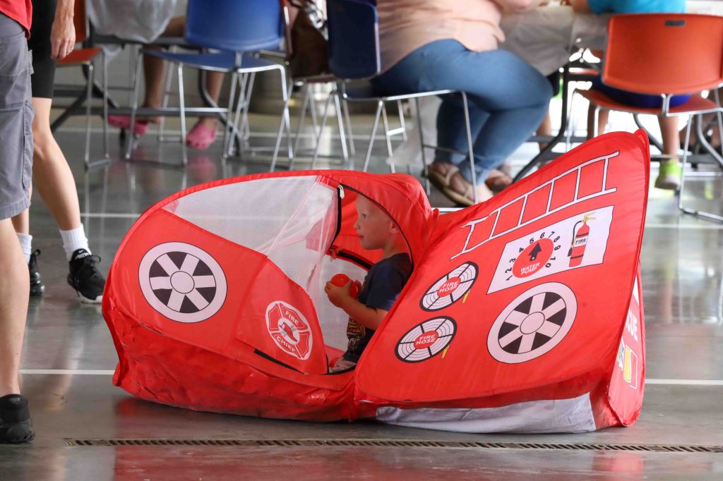 A young child playing inside a red fire truck-themed play tent on a smooth surface, with people seated in the background.