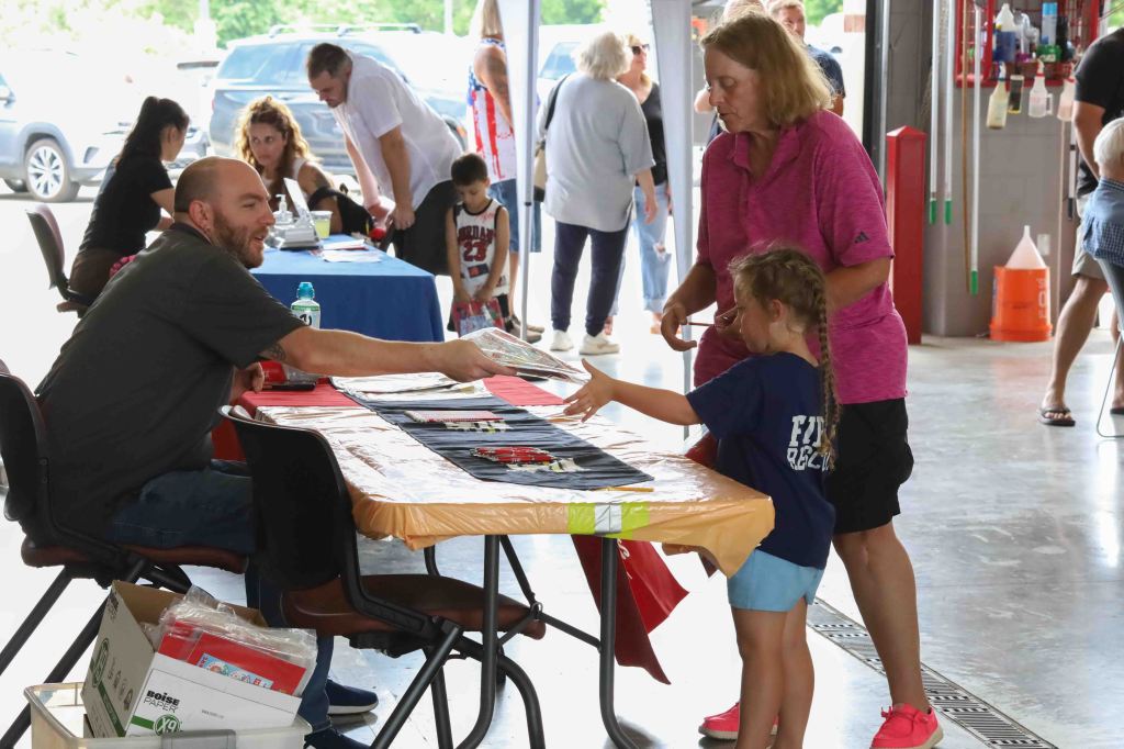 A vendor interacts with a child at a table while an adult stands beside them, both looking at materials laid out on the table. In the background, more people are engaged in activities at an indoor event.