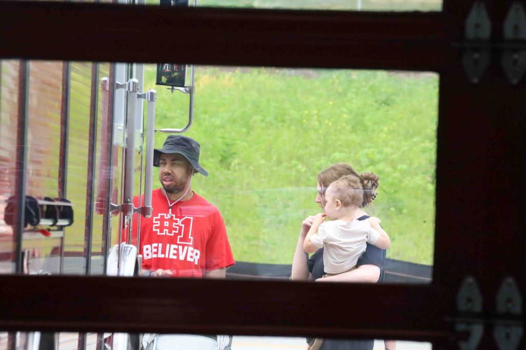 A man in a red '#1 BELIEVER' t-shirt and a black hat stands next to a truck, talking to a woman holding a toddler. The scene is set outdoors with greenery in the background.