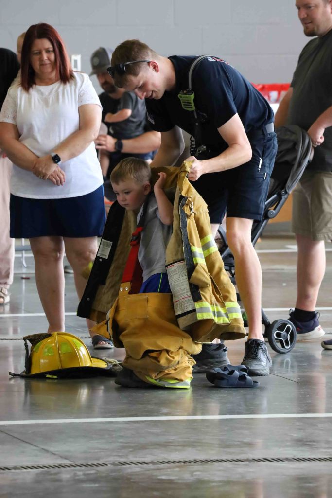 A firefighter helps a young boy try on a firefighter's jacket and pants while bystanders watch, including a woman and two men.